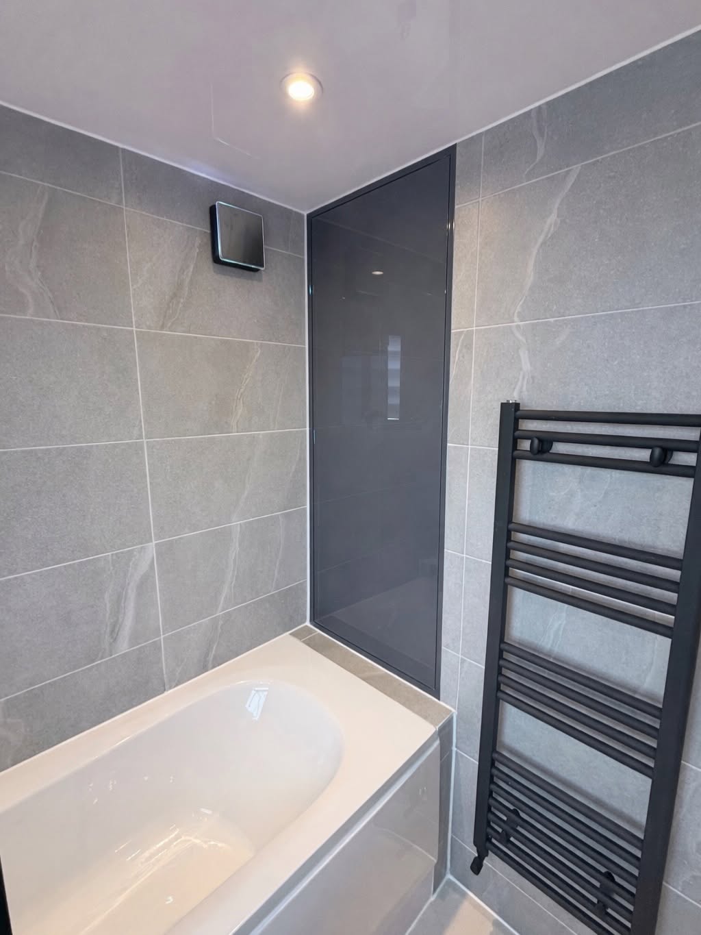 Grey tiled bathroom with bath and black heated towel rail